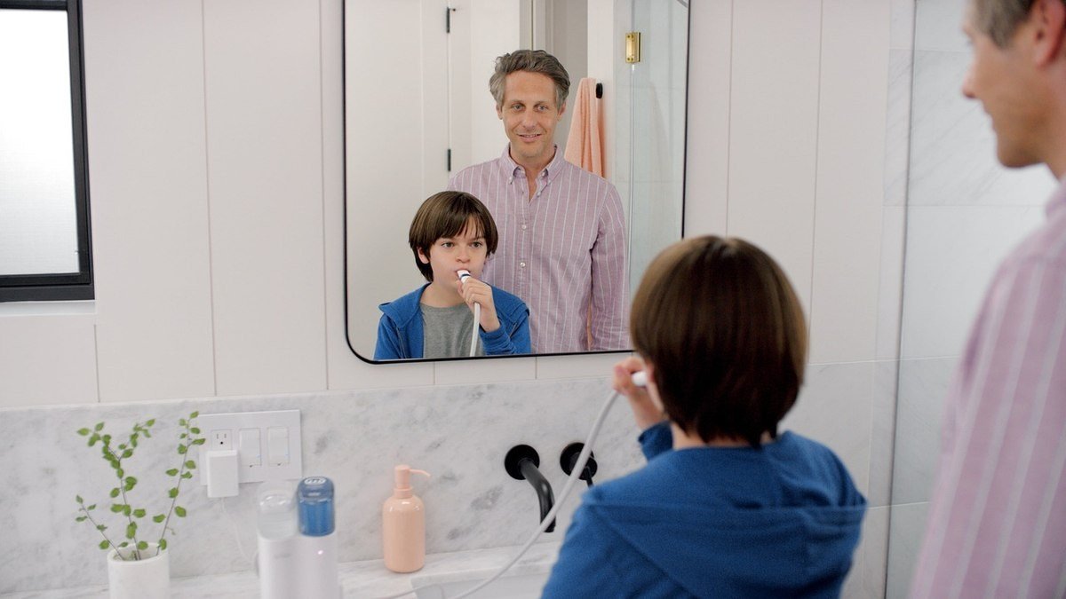 Enfant en bleu brossant ses dents dans une salle de bain moderne, parent observant derrière lui
