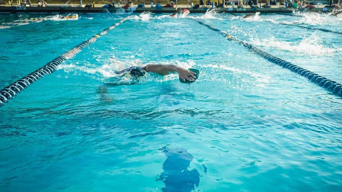 Nageur en bas-côté d'une piscine, bras tendus, eau bleue, cordes de séparation, autres nageurs en arrière-plan